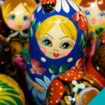 Links sind Schmetterlinge und das Logo der adi eingeblendet. In der Mitte sieht 7 russische Puppen in verschiedenen Größen, die eng nebeneinander stehen. In der Mitte ist eine größere Puppe mit blauem Kopftuch und gelben Haaren. Sie schaut aus dem Bild heraus. Die Gruppe ist von schräg oben fotografiert. Rechts ist das Logo vom Sommer der Vielfalt